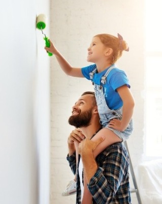 Girl sitting on father's shoulders painting a wall. 