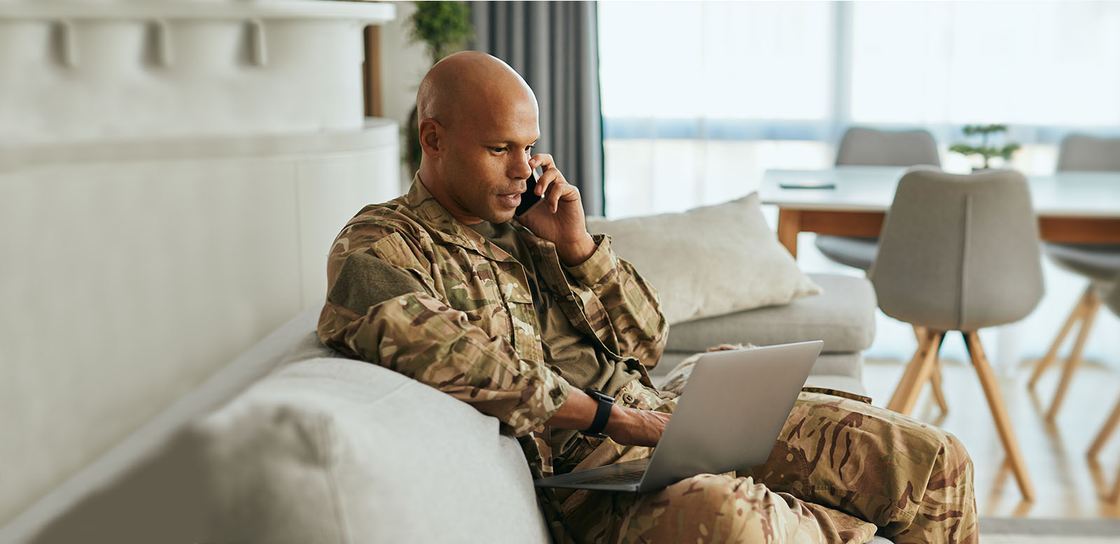 Male soldier in camouflage uniform sitting on the couch talking on a cell phone and looking at a laptop.
