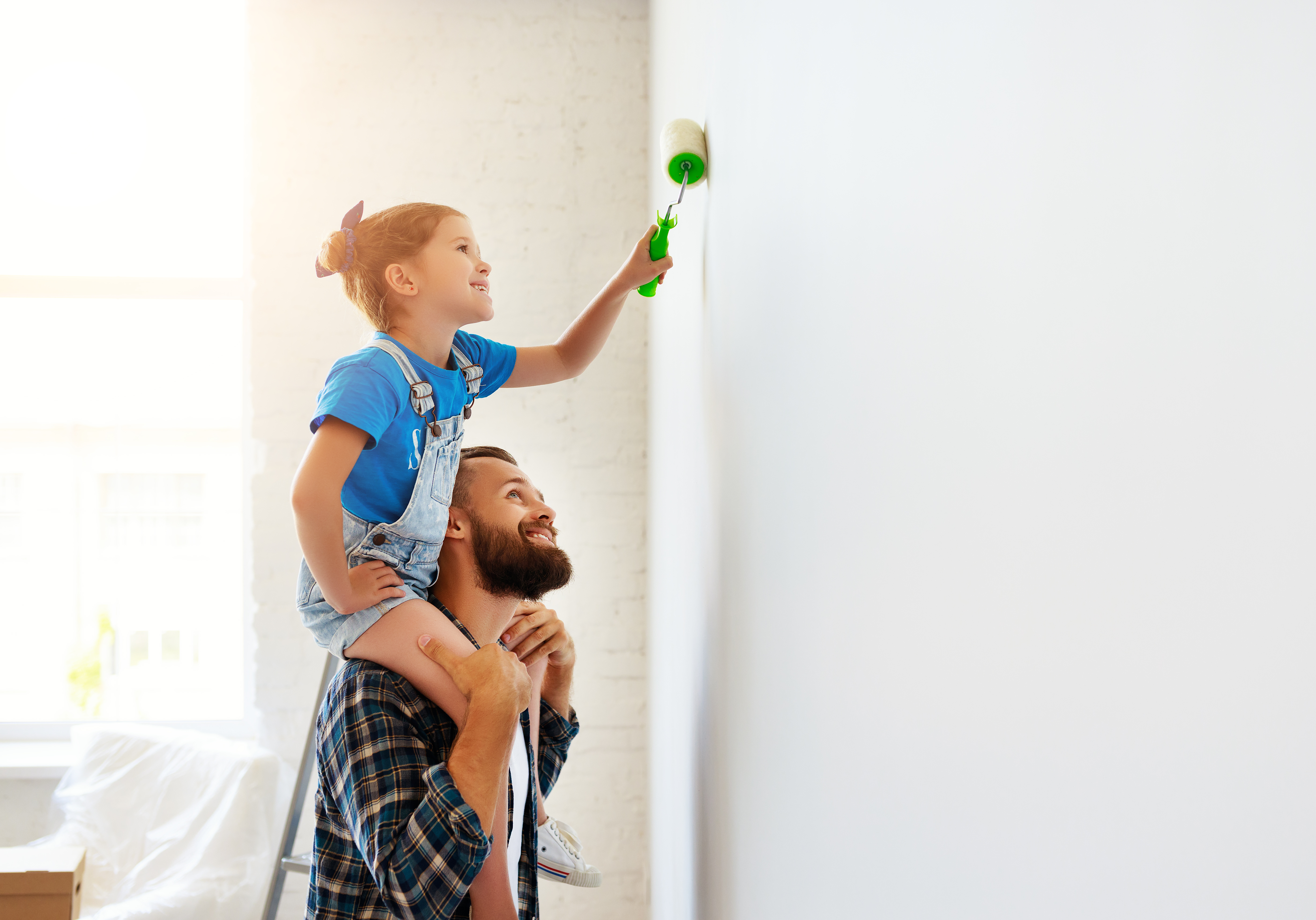 Girl sitting on father's shoulders painting a wall. 