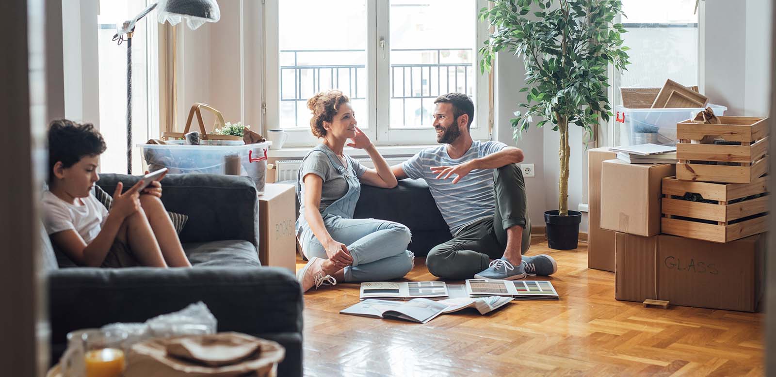A man and woman sitting on a floor talking with floor swatches on the floor. A kid is sitting on a couch with a phone.