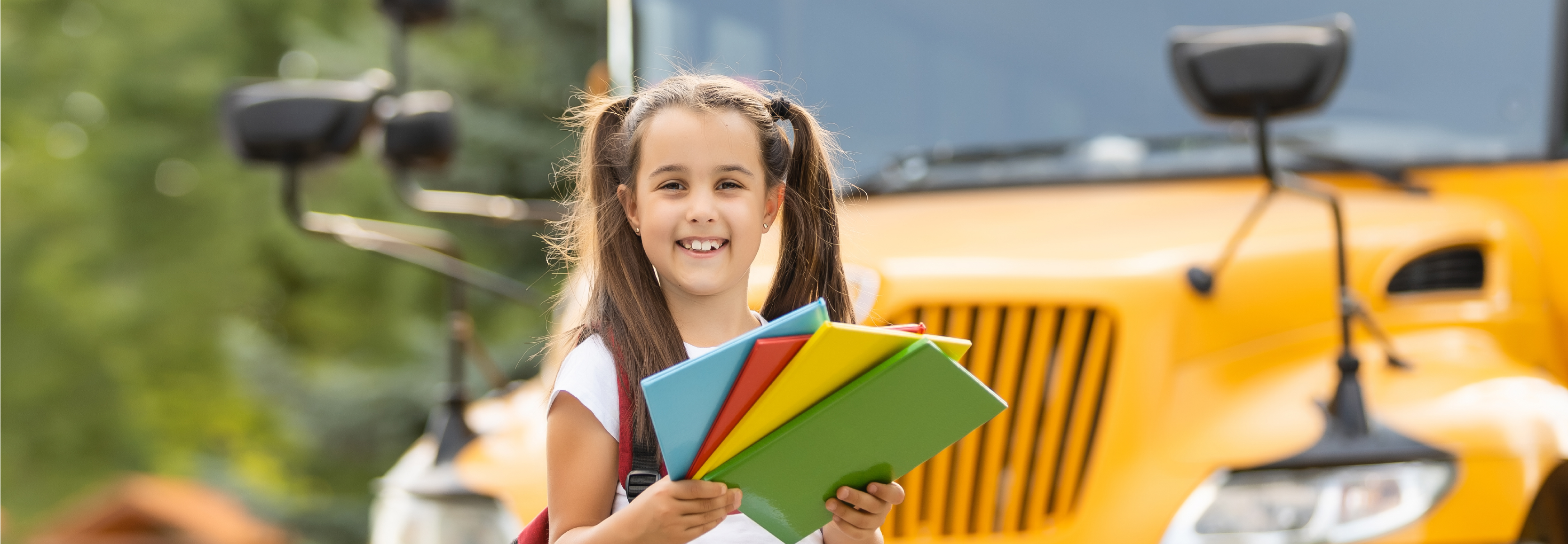 girl standing in front of school bus holding folders.