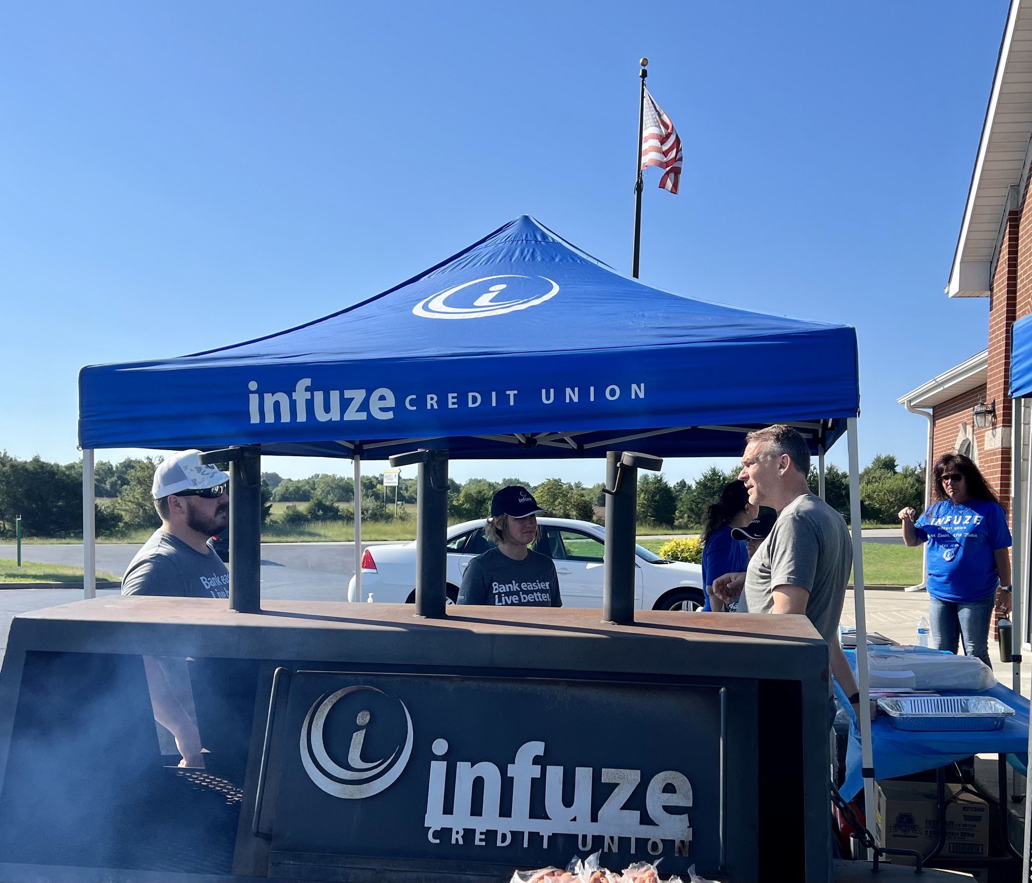 Group of people talking by a canopy and grill.