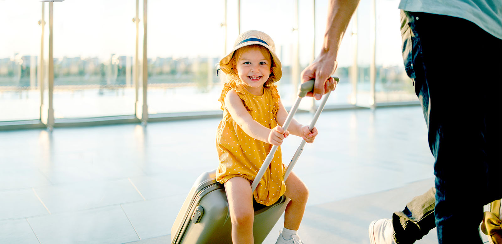 Young girl wearing yellow dress, sitting on top of a suitcase that is being pulled by a man.